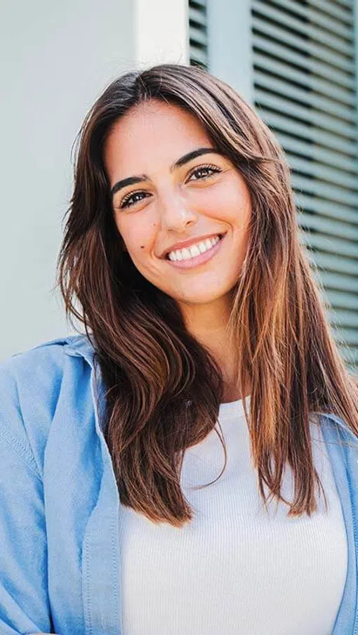 Smiling young woman with long hair wearing a blue shirt, standing in front of green shutters.