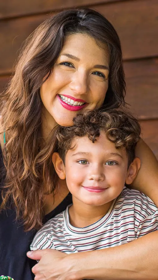Smiling woman hugging young boy with curly hair against wooden wall background