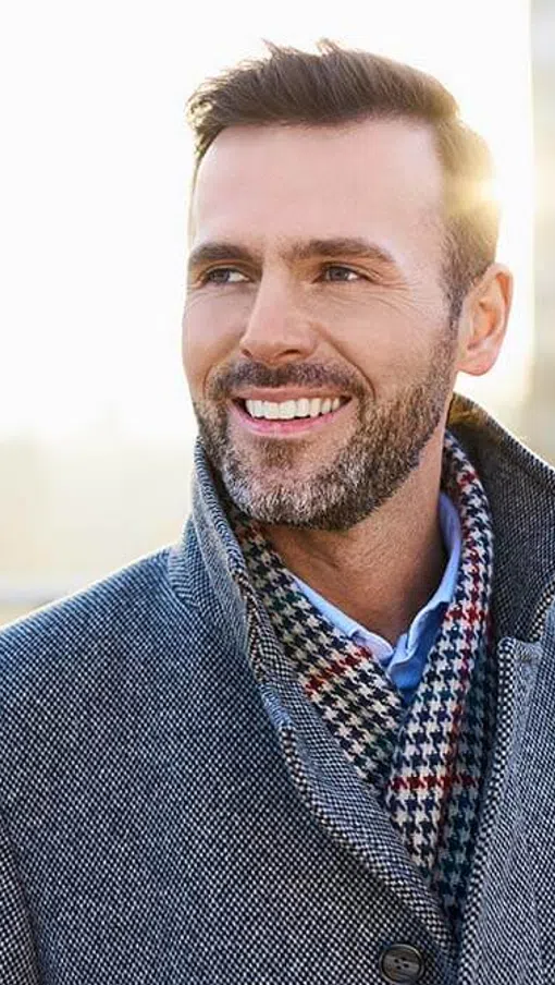 Smiling man in gray coat and patterned scarf enjoying a sunny day outdoors with blurred city background
