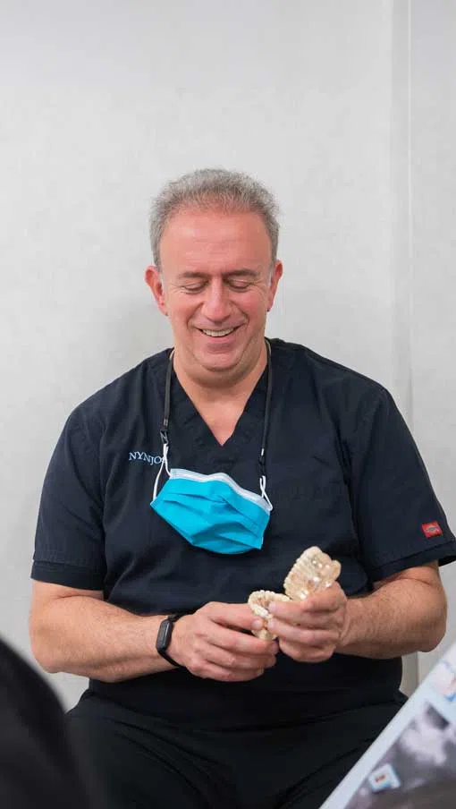 Smiling male dentist in black scrubs holding dental model with blue face mask around neck