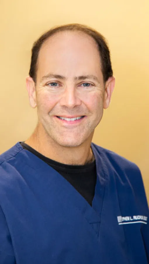 Smiling middle-aged man in blue medical scrubs with light brown background, facing the camera.