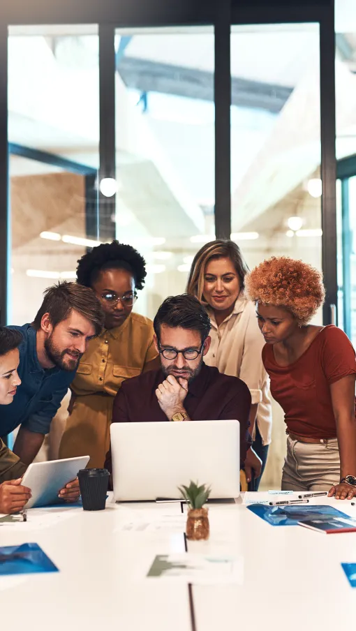 Diverse team gathered around a laptop in a modern office, collaborating and strategizing together.
