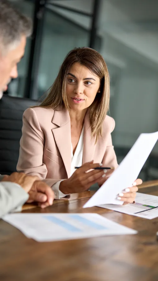 Businesswoman in a beige blazer discussing documents with a colleague at a wooden office table.