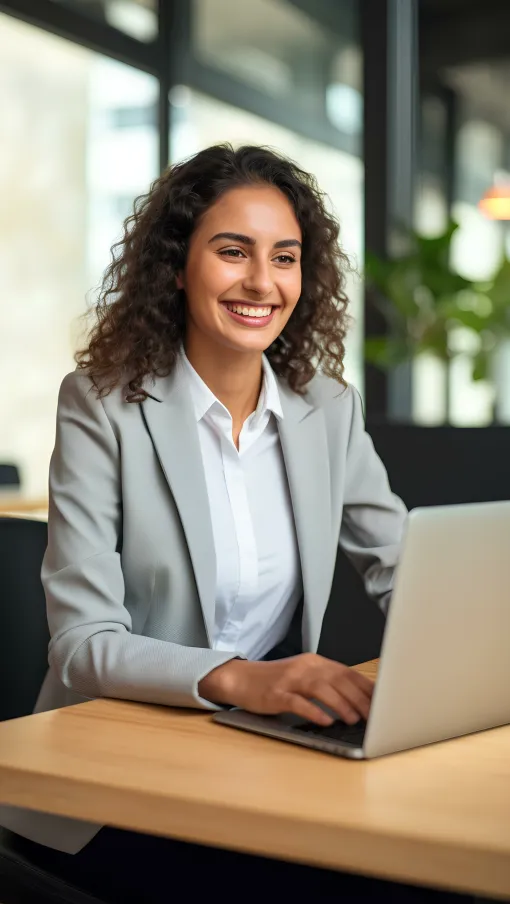 Smiling businesswoman in gray blazer working on laptop at modern office desk with blurred background.