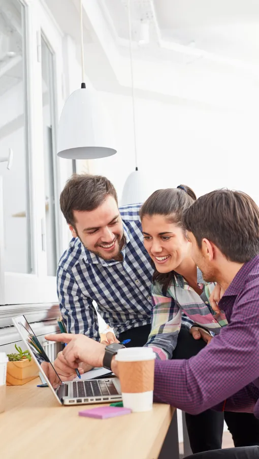 Three young professionals collaborating and smiling while discussing work on a laptop at a bright office desk.