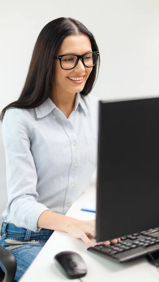 Smiling woman wearing glasses working on a desktop computer at a white office desk with a keyboard and mouse.