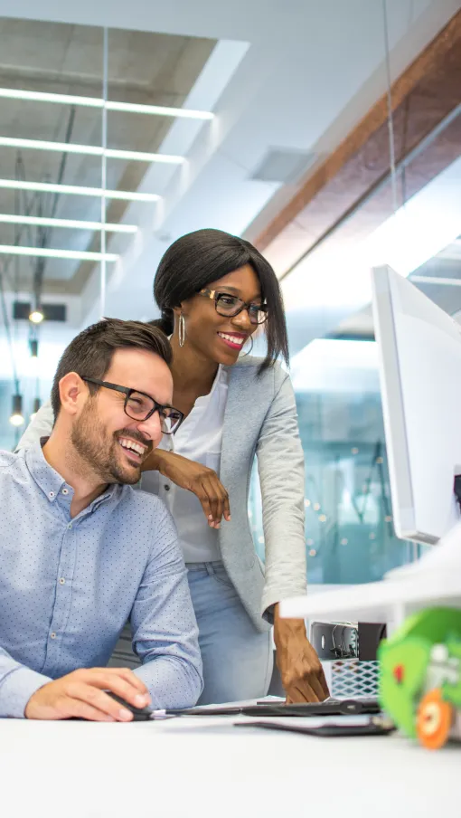 Two colleagues smiling and collaborating while working on a computer in a modern office space