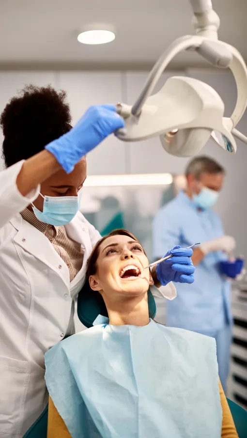 Dentist in blue gloves examining smiling patient's teeth with dental mirror and overhead light in clinic.