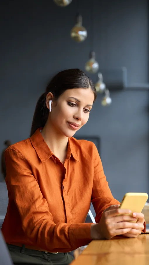 Young woman in orange shirt using smartphone with laptop nearby in a modern workspace by large windows.