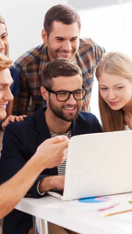 Group of five young adults smiling and collaborating while looking at a laptop screen in a bright office.