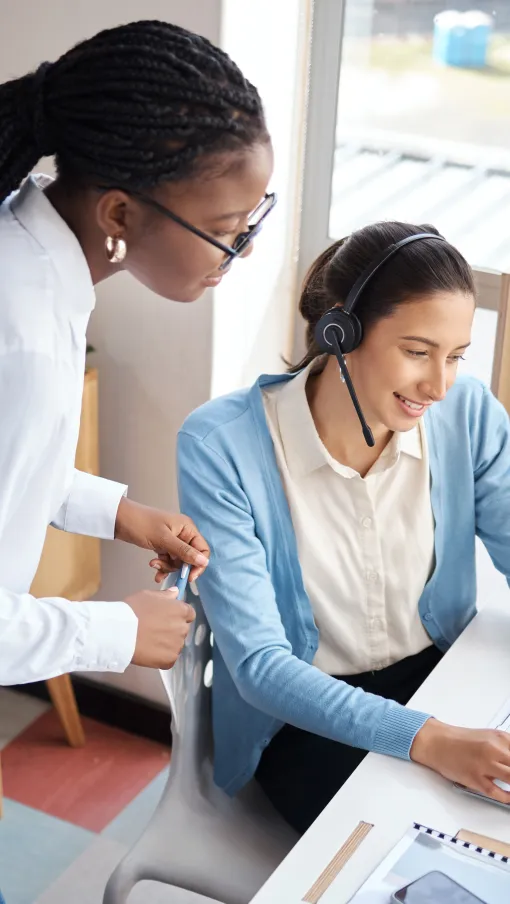 Two women collaborating in an office, one assisting with a laptop while wearing a headset.