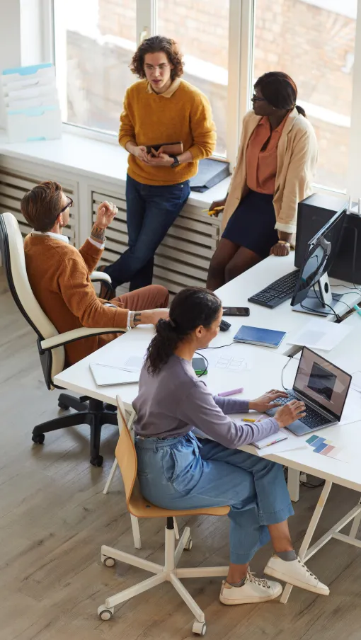 Four diverse colleagues engaged in discussion and laptop work in a bright modern office with large windows.