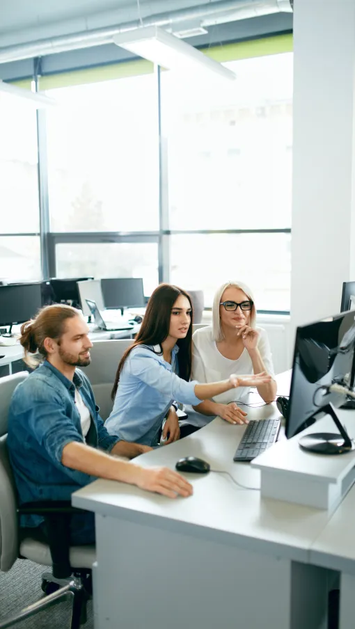 Three coworkers collaborating and discussing work on a desktop computer in a modern office.