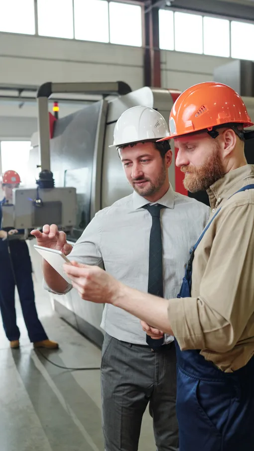 Engineer and technician wearing helmets reviewing plans on a tablet in an industrial factory setting.