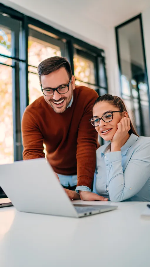 Smiling man and woman working together on a laptop in a bright modern office with large windows.