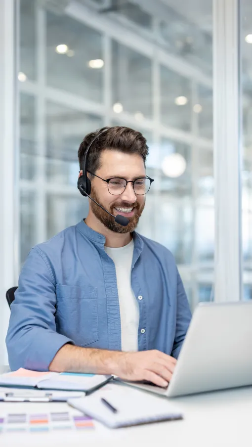 Smiling man wearing headset and glasses working on laptop in modern office environment.