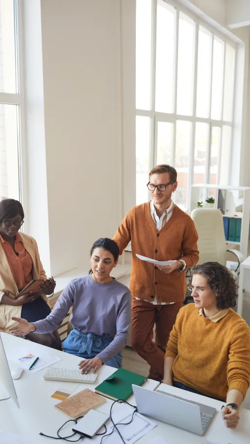 Diverse team collaborating in a bright office, discussing work at computer and laptops around a desk.