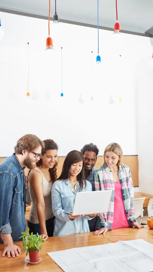 Group of diverse young adults collaborating and smiling while looking at a laptop in a bright office.