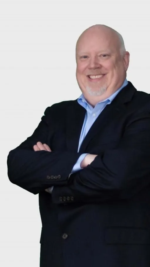 Smiling bald man in a blue shirt and dark blazer with arms crossed on a plain light background