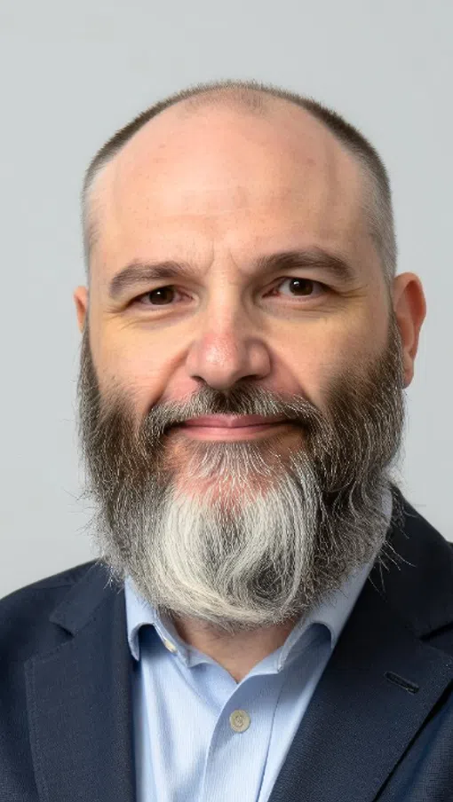 Middle-aged man with a full gray beard and bald head wearing a navy blazer and light blue shirt
