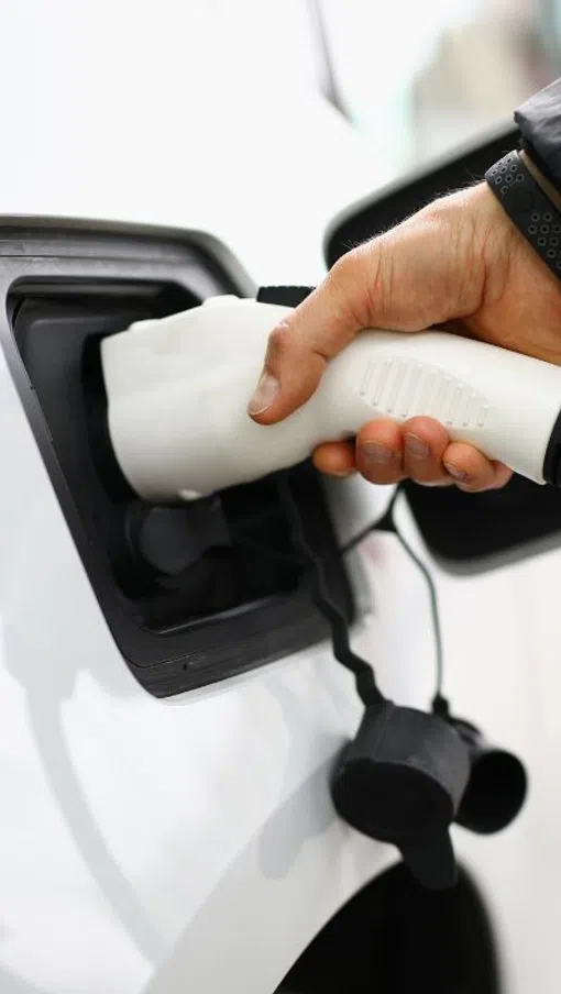 Person plugging a white electric vehicle charger into a white car at a charging station outdoors.