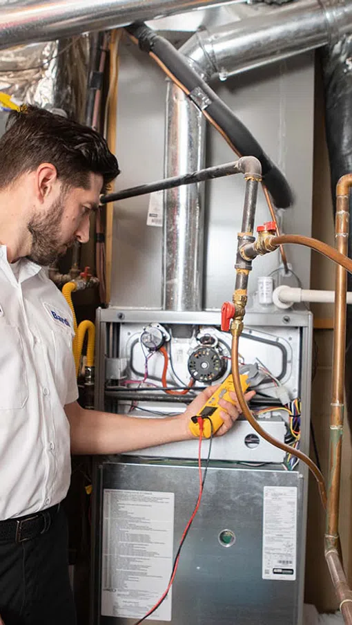 Technician inspecting a heating system with tools in a mechanical room full of pipes and equipment.
