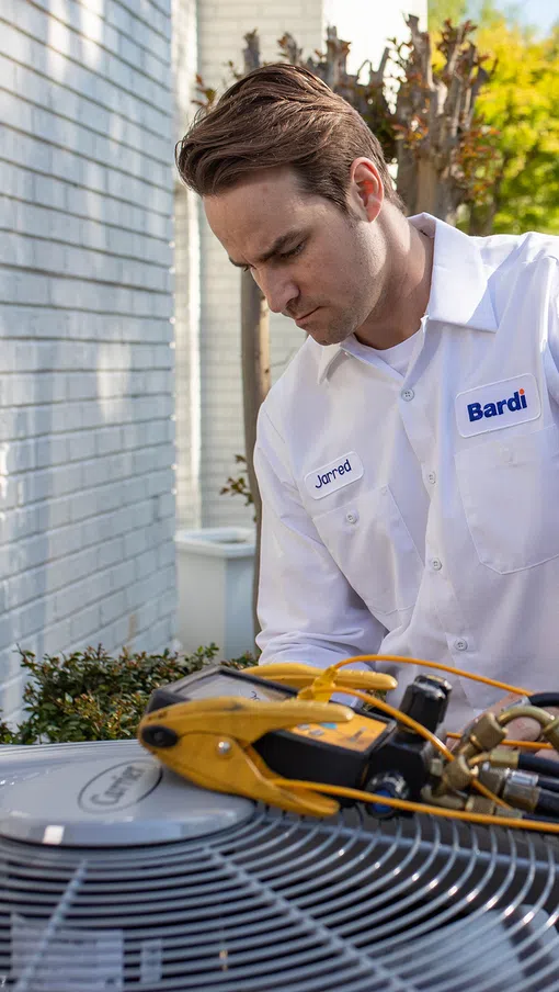 HVAC technician servicing an outdoor HVAC unit with a Bardi service van in the background.