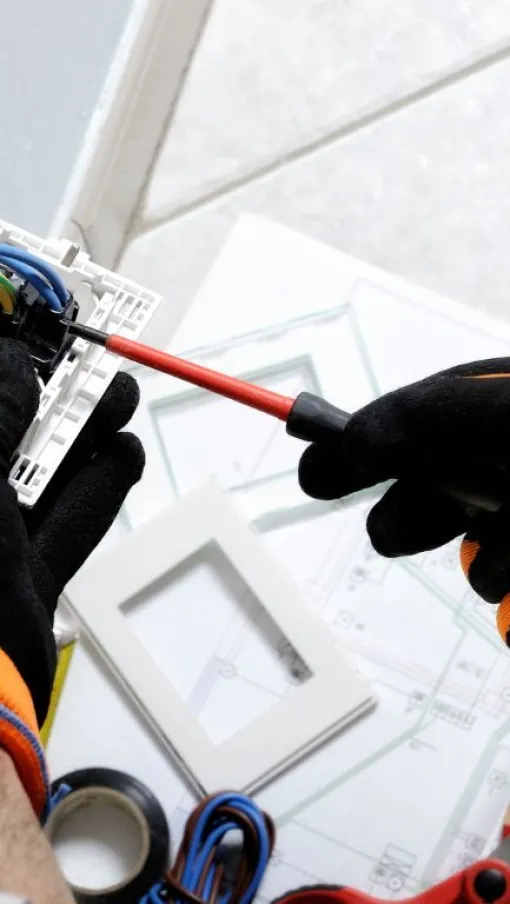 Electrician wearing orange gloves installing a wall switch with wires and tools on a floor blueprint