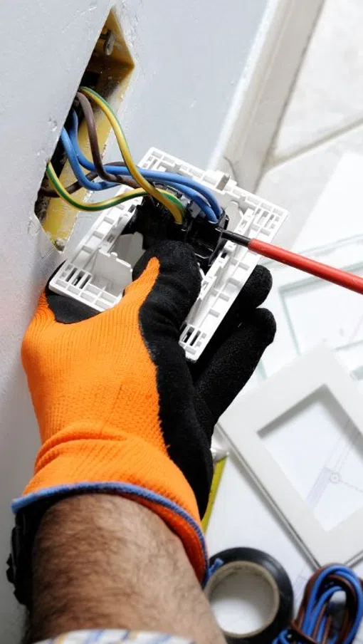 Electrician wearing orange gloves installing a wall switch with wires and tools on a floor blueprint