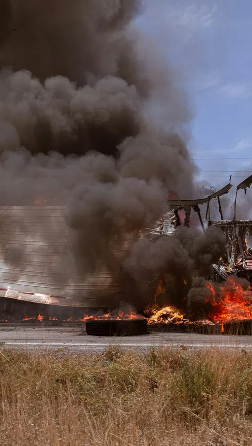 Truck trailer engulfed in flames with thick black smoke rising on an open highway near a kilometer road sign.