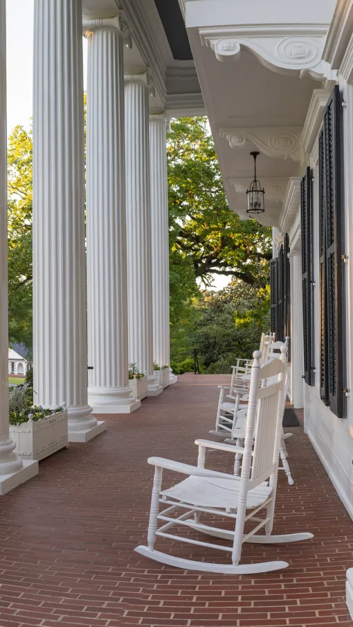 Wide porch with white columns and rocking chairs overlooks green lawn and trees at sunset.