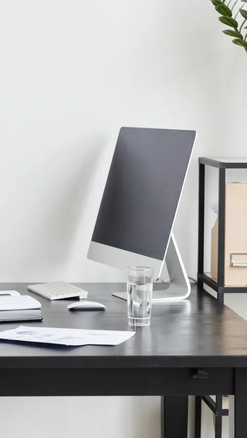 Modern minimalist office workspace with computer, glass of water, notebook, and plants on black desk with chair.