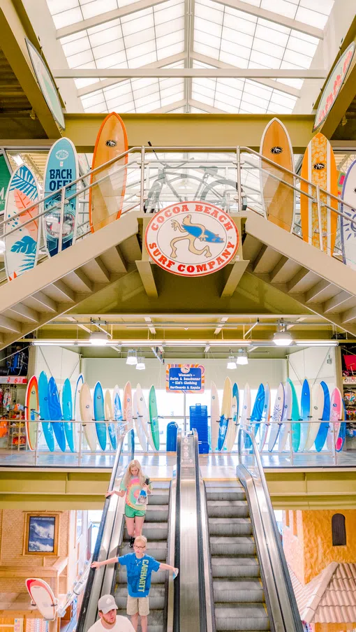 Interior of a surf shop with colorful surfboards displayed around a staircase and two people on an escalator.