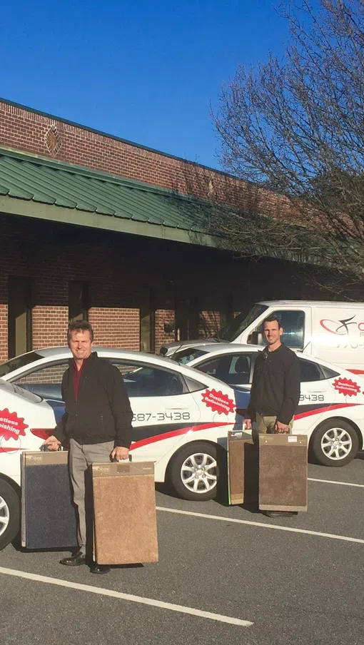 Three men holding carpet samples in front of three Concept Flooring company vehicles parked outside a brick building under a green roof.