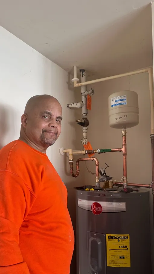 Smiling Estes Plumber technician in orange shirt standing beside a Rheem water heater with copper piping in a utility room.
