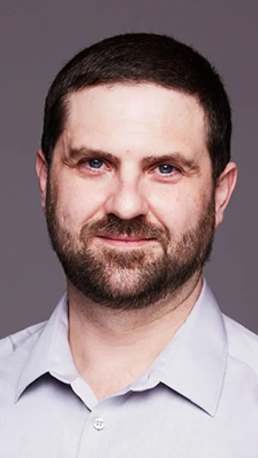 Headshot of a man with short dark hair and beard wearing a light gray collared shirt smiling against a plain gray background