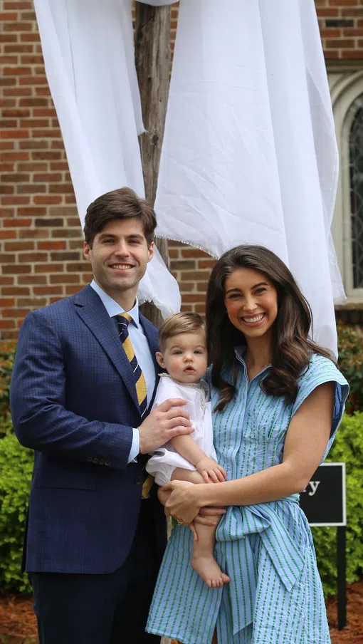 Young family with father, mother, and baby posing outside a brick building with greenery and white drapes.