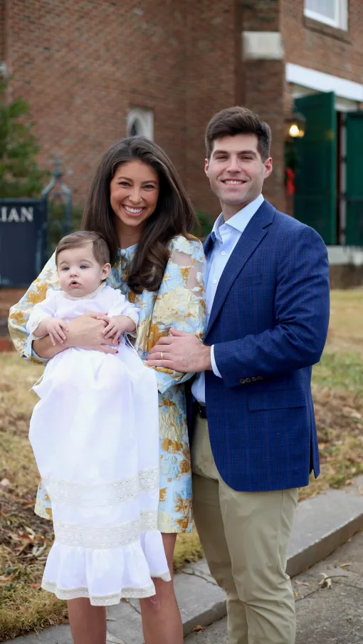 Smiling couple holding a baby in a white gown outdoors near a church building with a sign and green door