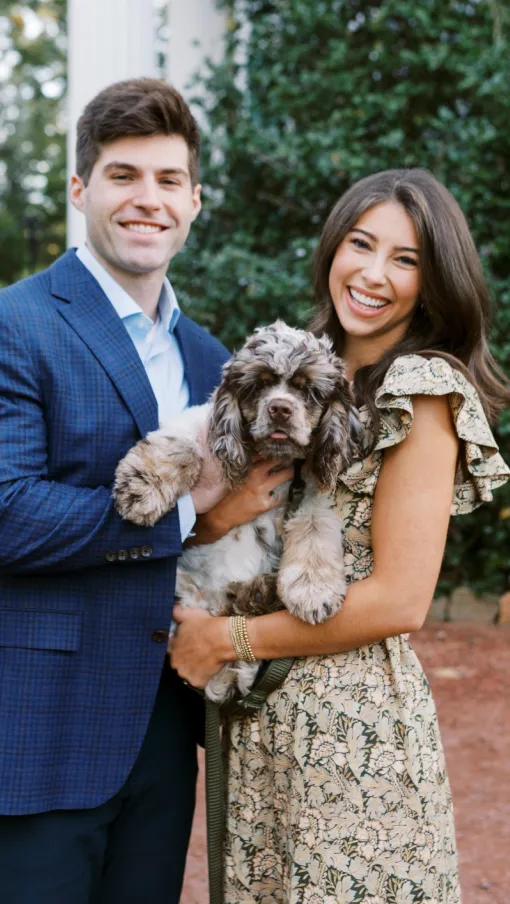 Smiling couple holding a dog outdoors, surrounded by greenery and bright sunlight.