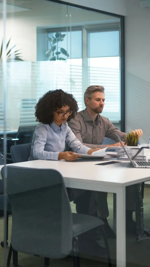 Three professionals collaborate around a white table in a modern glass-walled office room with laptops and documents.