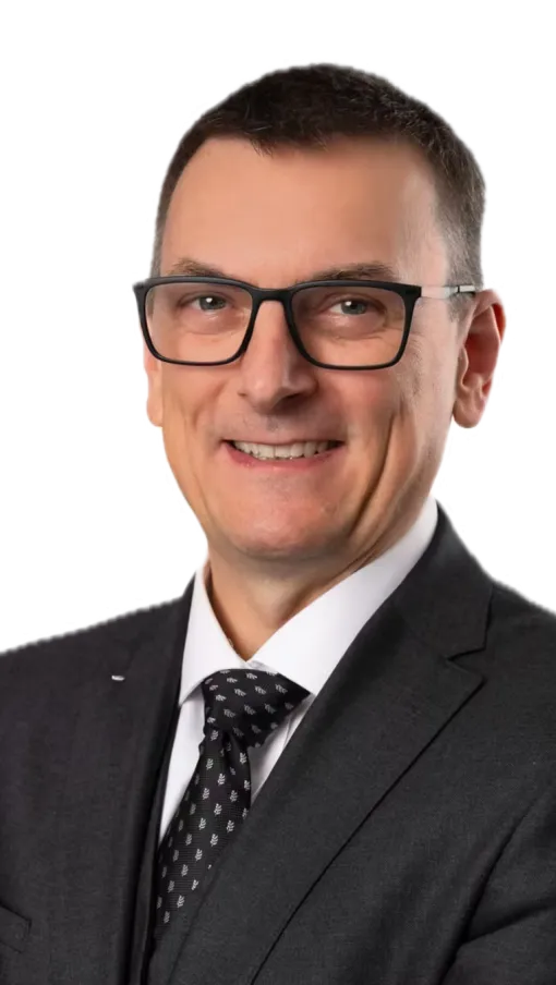 Smiling middle-aged man in black suit, patterned tie, and glasses posing with arms crossed on white background.