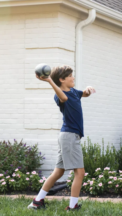 Boy in navy shirt playing catch with football in a garden in front of a white brick house.