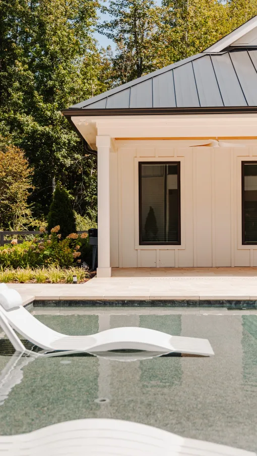 Modern poolside lounge chair in shallow water near a light-colored house with a metal roof and garden trees.