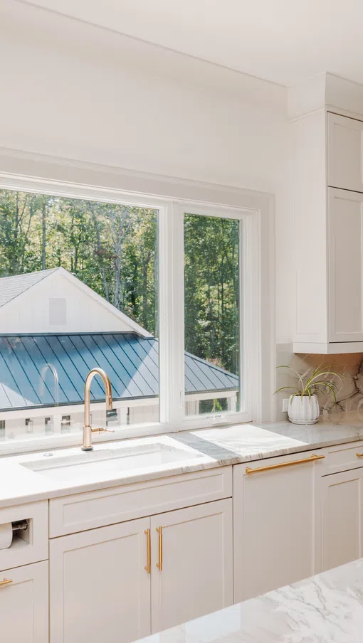 Bright modern kitchen with white cabinets, marble countertops, gold hardware, and large window overlooking trees.