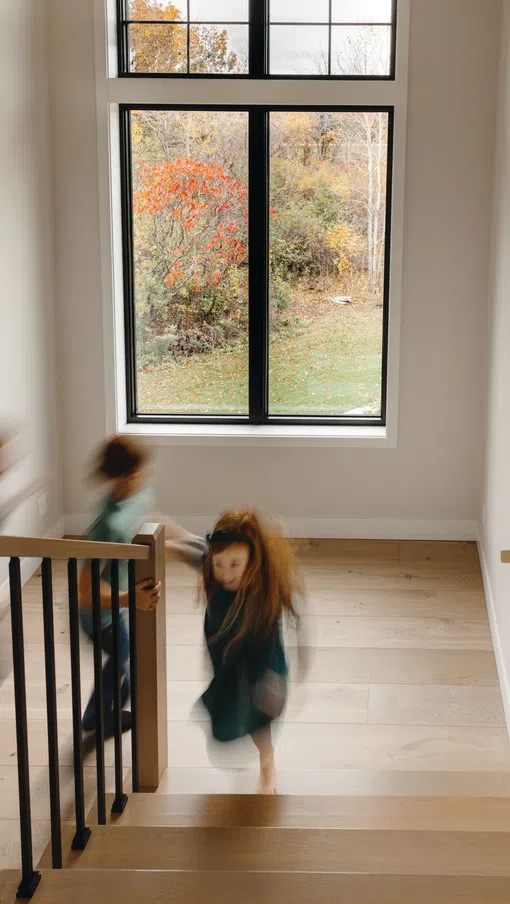 Children in motion running up a wooden staircase inside a bright home with a large window showing autumn trees outside.