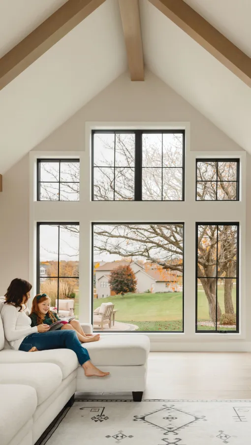 Mother and daughter reading on a white couch in a living room with large decorative windows and autumn view.