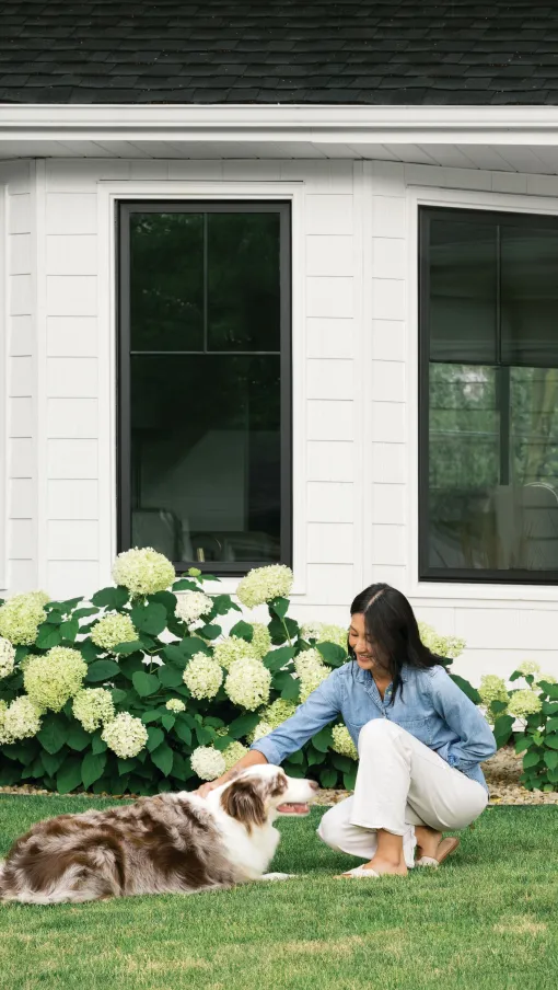 Woman in sunglasses watering flower garden with hose in front of white brick house on a sunny day