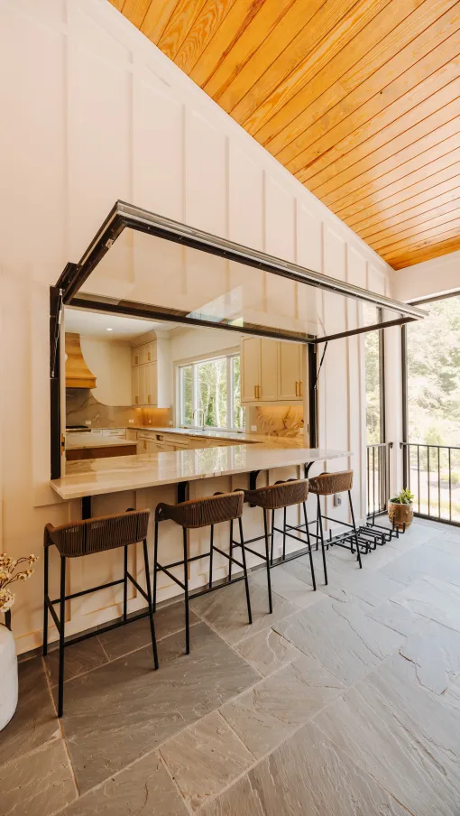 Open kitchen pass-through window with a marble bar counter and four bar stools on stone tile floor under a wooden ceiling.
