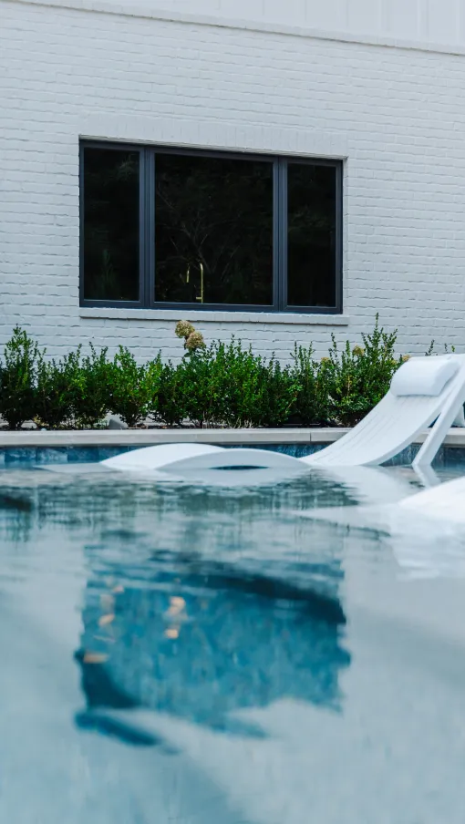 White lounge chairs partially submerged in a backyard pool in front of a white brick house with greenery.