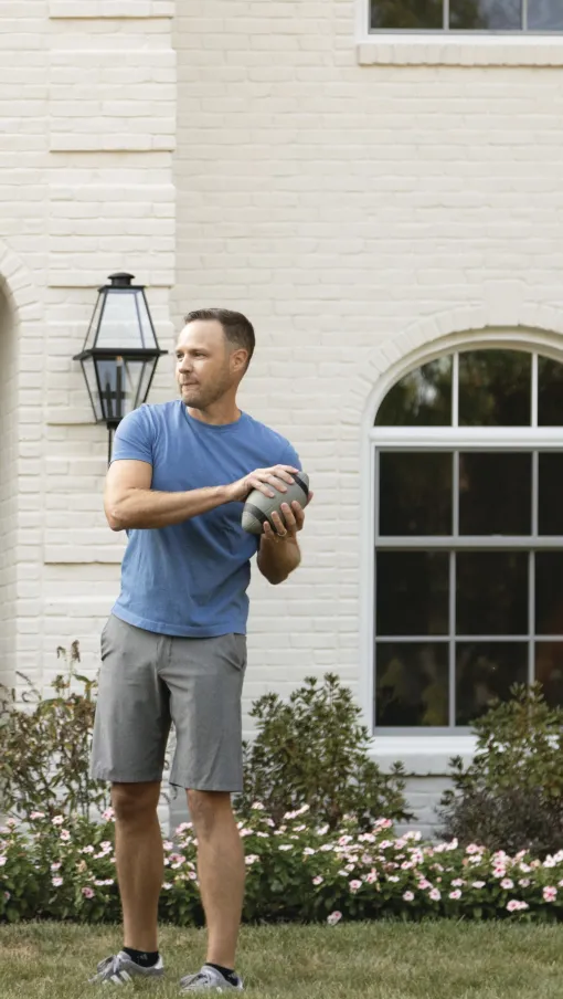 Man in blue shirt and gray shorts holding football, preparing to throw in front of white brick house with garden.